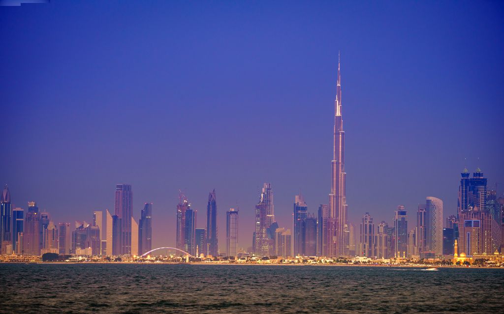 Skyline of Dubai at twilight with the Burj Khalifa towering over the city, its silhouette piercing the dusky sky, surrounded by the illuminated cityscape and reflected on the tranquil waters in the foreground.
