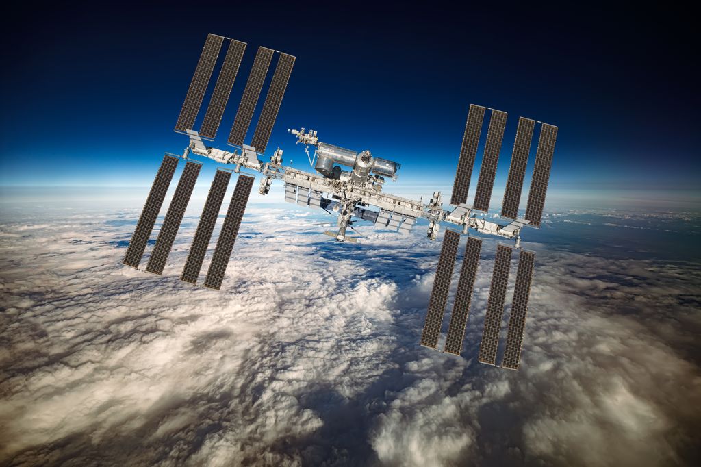 The International Space Station (ISS) orbits Earth, with its expansive solar arrays fully extended against a backdrop of the planet's atmosphere and a blanket of clouds below.