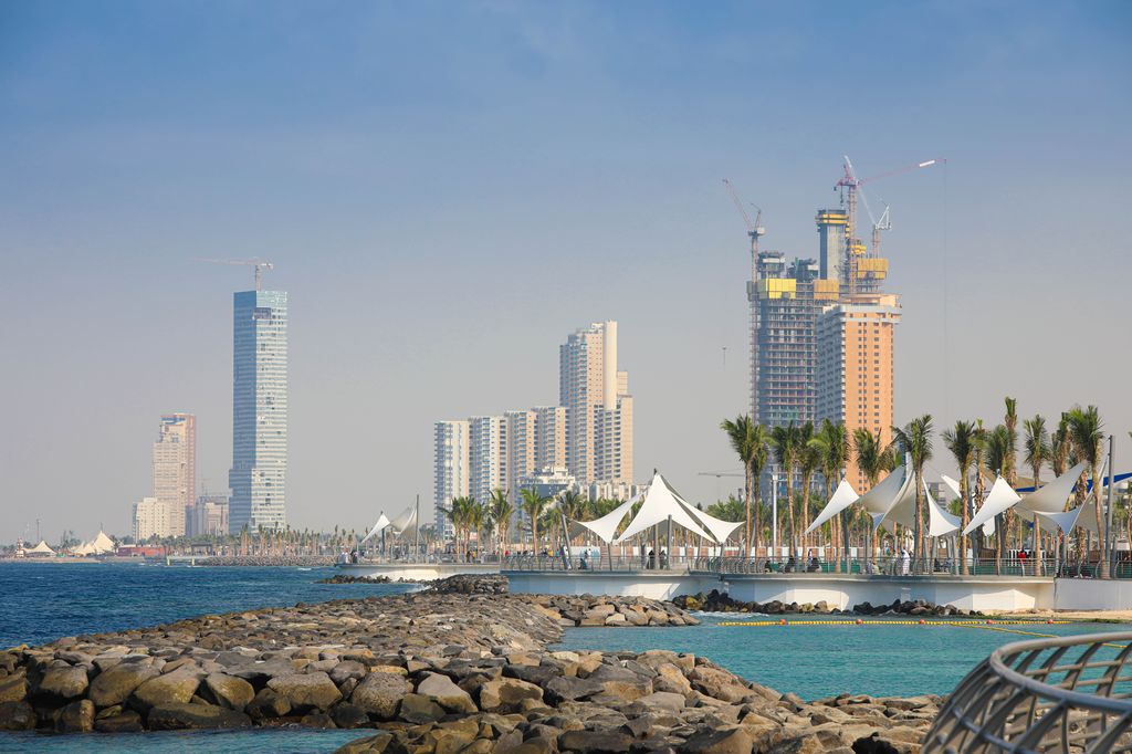 View of Jeddah's waterfront with modern high-rise buildings and construction cranes against a clear sky, and the Jeddah Tower under construction in the distance, all seen from a rocky shoreline with distinctive white sunshades along the promenade.
