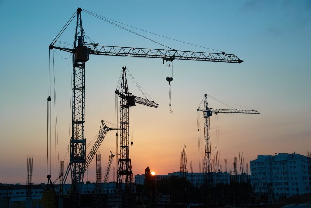 Silhouette of construction cranes at a building site against a sunset sky, with the sun dipping just below the horizon, reflecting the progress of one of the world's largest construction projects.