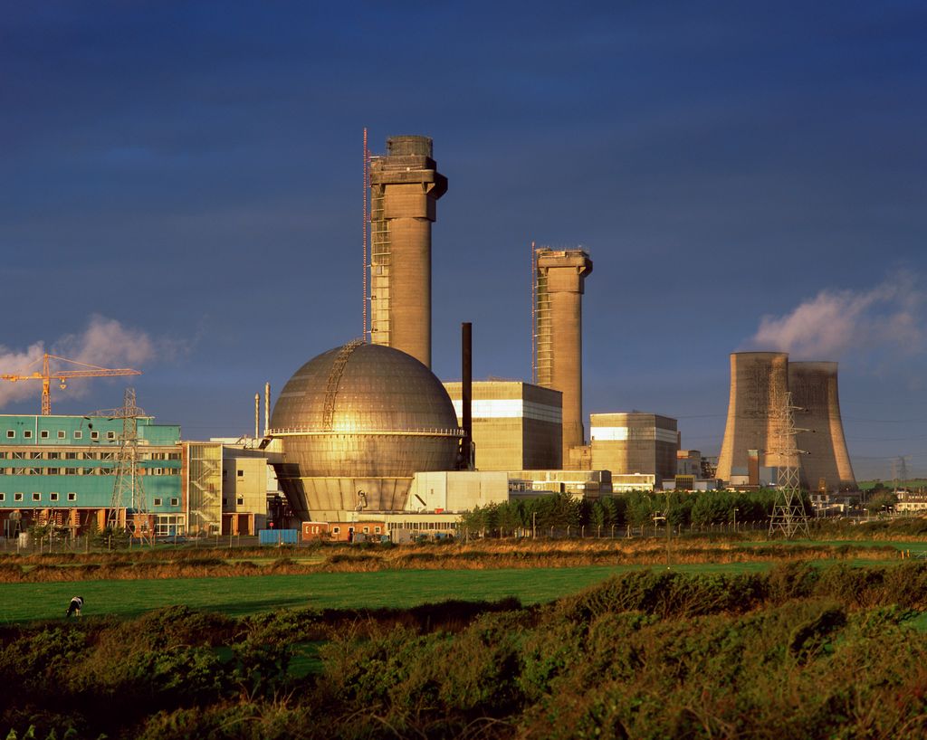 The Sellafield nuclear reprocessing facility under a clear blue sky, with its distinctive dome-shaped reactor building, tall cooling towers emitting steam, and ongoing construction indicated by cranes, set against a lush green field.