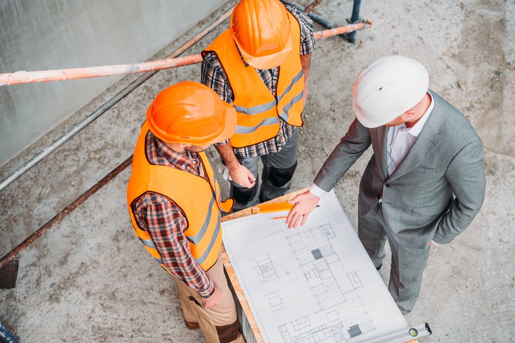 Two construction workers and an architect in a business suit, all wearing safety helmets, are examining blueprints on a work site, highlighting a moment of professional consultation and planning in the construction lifecycle.