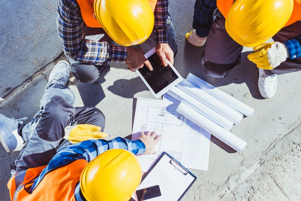 A top-down view of a construction team huddled over architectural plans on a concrete surface, with several individuals wearing high-visibility vests and hard hats, holding rolled-up blueprints and a tablet, illustrating a collaborative outdoor work meeting.