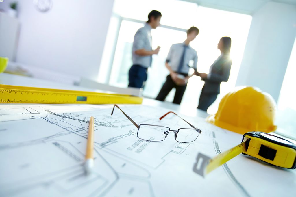 A close-up of architectural blueprints with a yellow ruler and glasses in focus, while in the background, out of focus, three professionals are engaged in discussion near a bright window, symbolizing a planning meeting in an architectural or construction setting.