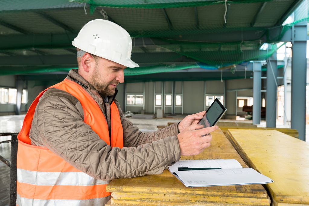 A construction worker wearing a white hard hat and orange safety vest is using a tablet while reviewing blueprints at a construction site.