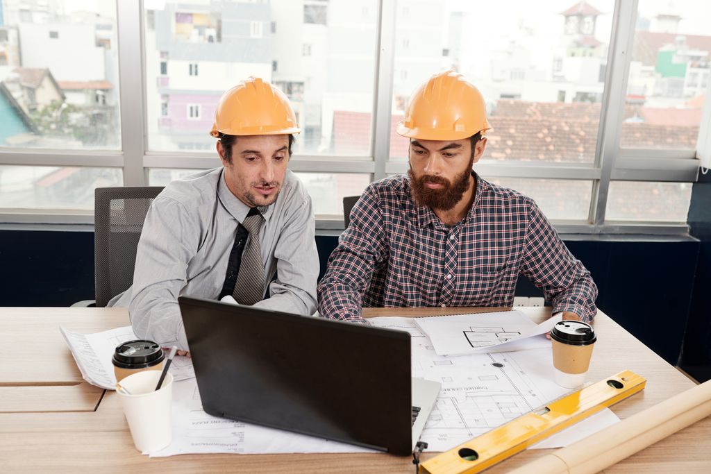 Two construction professionals wearing yellow hard hats are working together at a desk with a laptop, blueprints, and coffee cups. This image represents project management using the secure construction cloud.