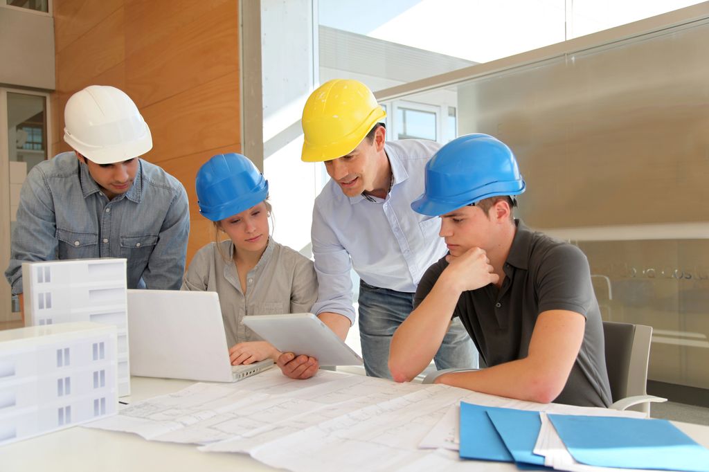 A group of construction professionals wearing hard hats are gathered around a table with blueprints, a laptop, and a tablet, actively collaborating on a project. This image represents team involvement and the integration of project planning and management.