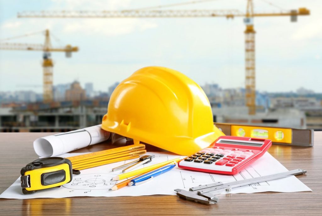 Construction tools and safety gear on a desk with cranes in the background, representing various phases of construction.